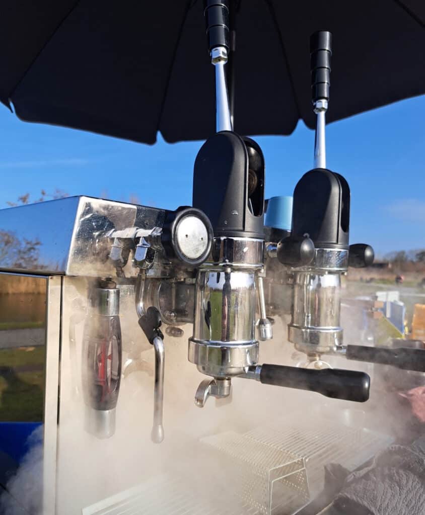 Professioneel koffiezetapparaat voor outdoor gebruik tijdens trainingen en bootcamps, onder een grote parasol met een heldere blauwe lucht op de achtergrond.