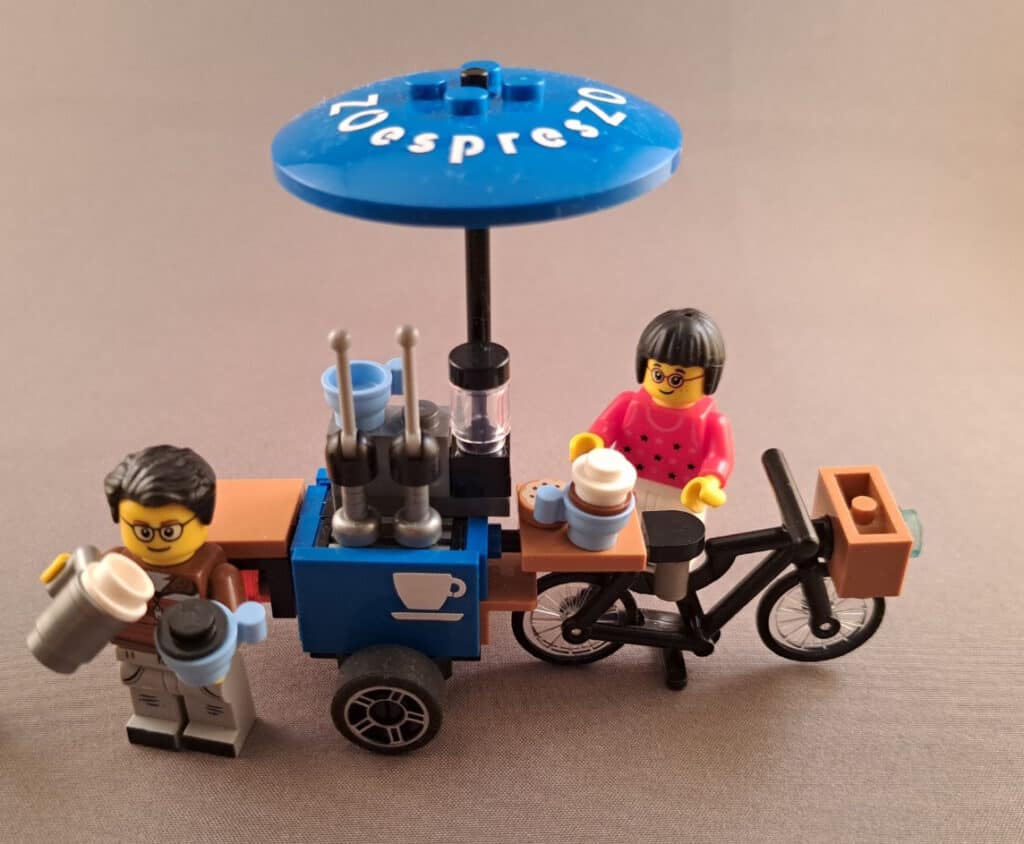 LEGO-beeld van een mobiele koffiebar met twee mensen, een op de fiets en een bij de koffie, tijdens een training of evenement.